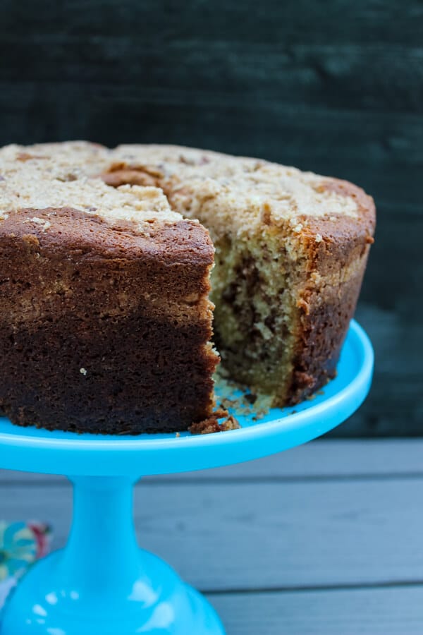 Side view of sour cream coffee cake with toasted pecan filling, set on a blue cake stand. Cake has a slice taken out of it, showing off the pecan swirl