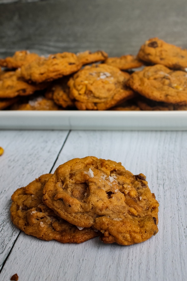 Salted Butterscotch Pretzel Cookies piled on a white plate in the background with two cookies in the foreground, against a white background. 