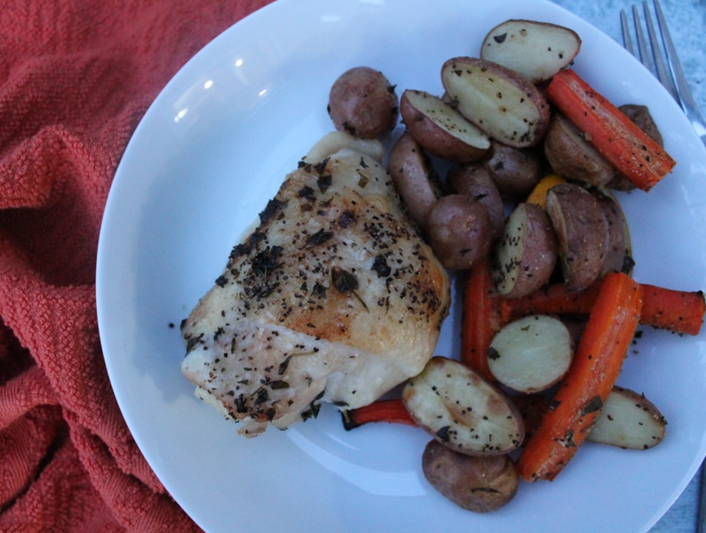 Overhead shot of roasted chicken thigh, potatoes and carrots in a white bowl, with an orange background