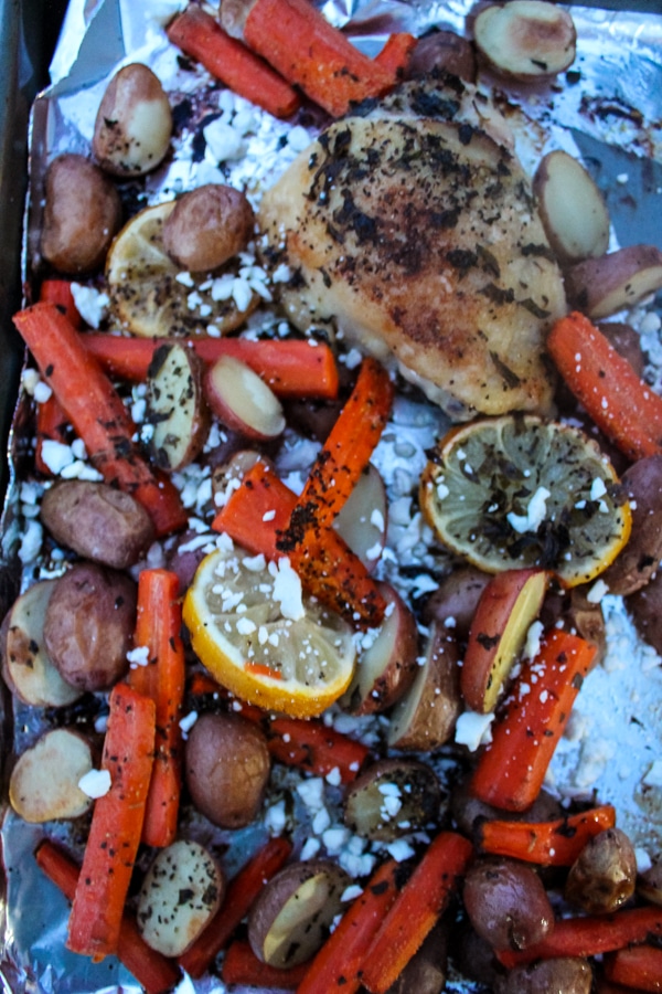 Overhead shot of roasted chicken, potato, carrot on a foil-covered sheet pan, sprinkled with crumbled white feta cheese