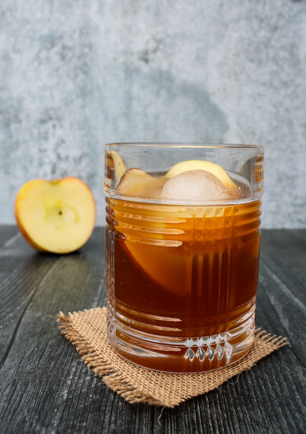 Side view of apple cider old fashioned. Glass set on a square of brown burlap on a black background, with half an apple in the background