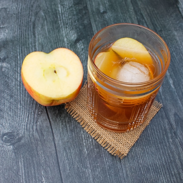 Overhead view of apple cider old fashioned. Glass set on a square of brown burlap on a black background, with half an apple next to the glass