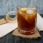 Side view of apple cider old fashioned. Glass set on a square of brown burlap on a black background, with half an apple in the background