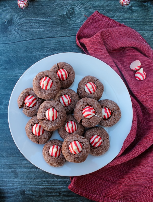 Overhead view of chocolate cookies with candy cane kisses on a white plate. The plate is set on a red cloth on a black background