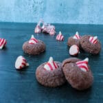side view of chocolate cookies topped with a candy cane kiss on a black background, unwrapped kisses scattered among the cookies
