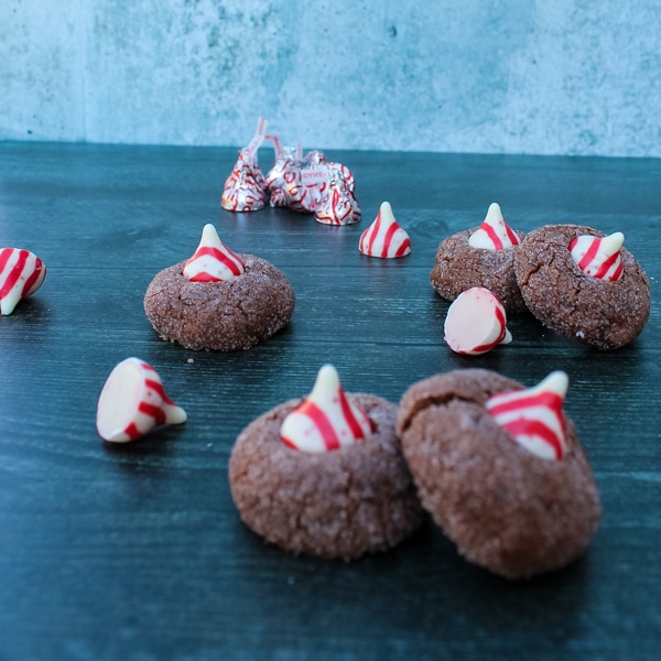 side view of chocolate cookies topped with a candy cane kiss on a black background, unwrapped kisses scattered among the cookies