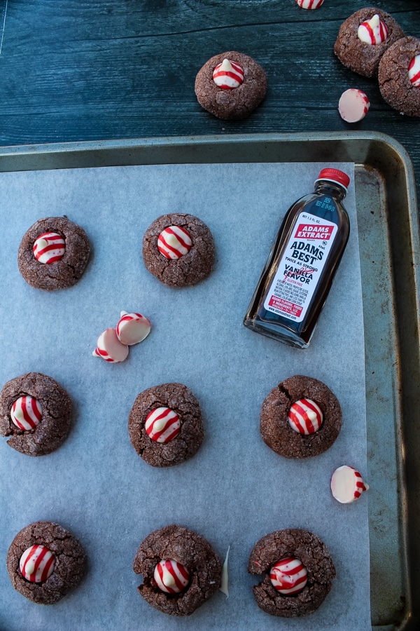 overhead view of chocolate cookies with candy cane kisses and a bottle of adams vanilla extract on a parchment lined baking sheet