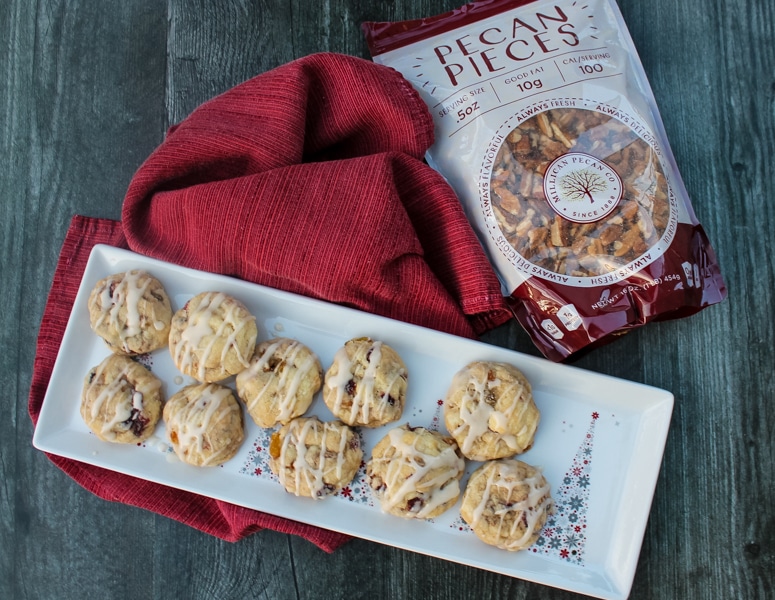 Overhead view of fruitcake cookies with rum glaze. Cookies sitting on a white plate. Plate of cookies and bag of pecans are sitting on a red napkin and a black background