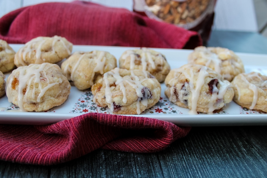 Side view of fruitcake cookies with rum glaze on a white plate, set on a red napkin on a black background. Bag of pecans peeking out in the upper right corner