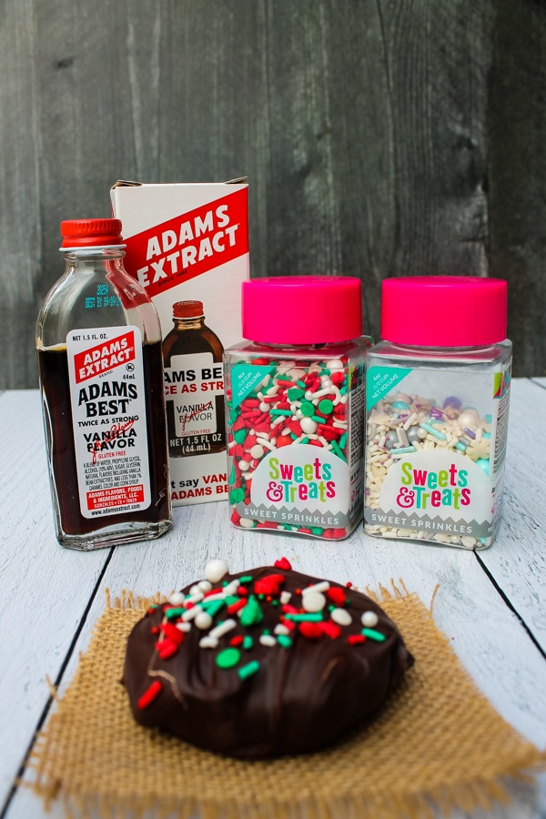 side view of a chocolate covered peanut butter cookie set on a piece of brown burlap on a white background with ingredients (vanilla extract and sprinkles) in the back, set on a black background