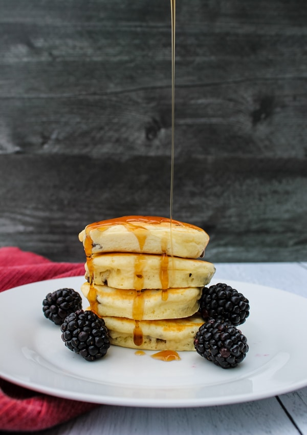 side view, stack of pancakes and a few blackberries on a white plate, with syrup being drizzled