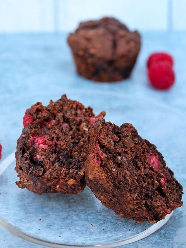 half a chocolate raspberry muffin on a plate in foreground, a muffin and a few raspberries in background