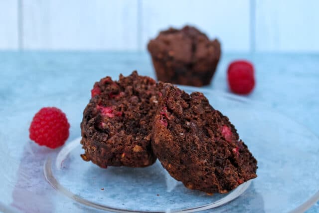 half a chocolate raspberry muffin on a plate in foreground, a muffin and a few raspberries in background