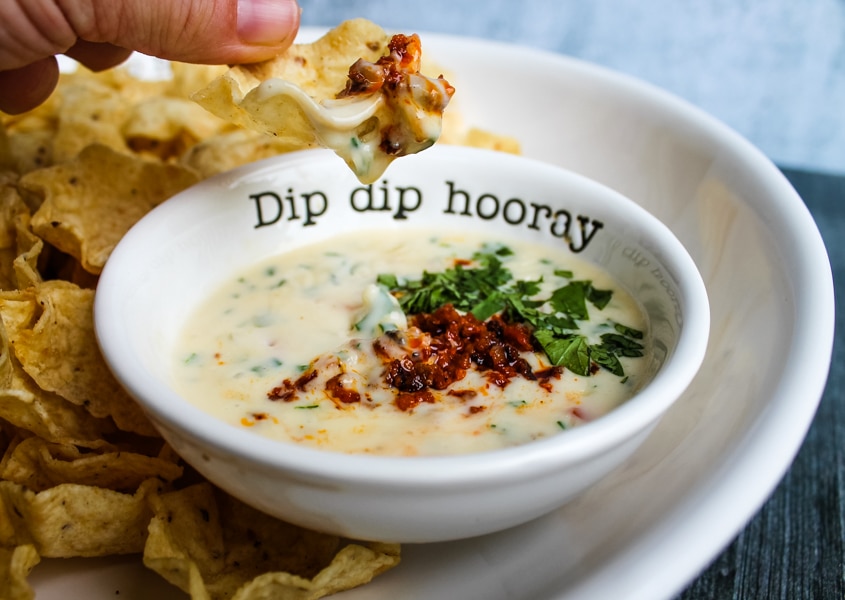 side view of a tortilla chip being dipped into a bowl of queso topped with chorizo. platter of chips and queso in background