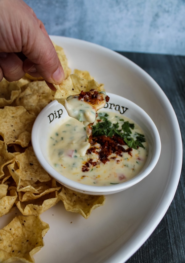 side view of a tortilla chip being dipped into a bowl of queso topped with chorizo. platter of chips and queso in background