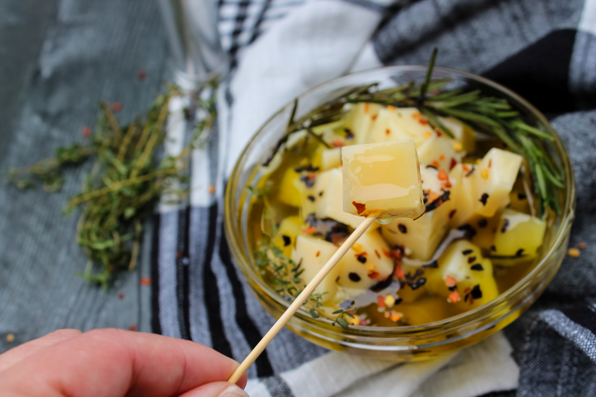 overhead view of Manchego Marinated in Olive Oil & Herbs on a toothpick over clear bowl