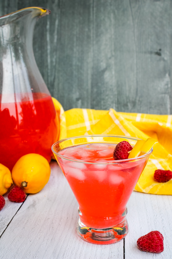 Side view of a glass of Raspberry Lemonade Limoncello Cocktail with a pitcher of the cocktail in the background